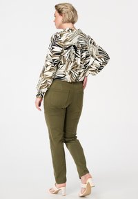 Femme avec des cheveux blonds courts portant un chemisier à motif feuillu, un pantalon vert olive et des sandales à talons beiges, se tenant avec une main sur la hanche.
