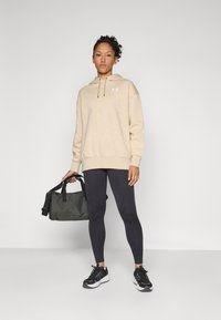 Beige hoodie med dragsnören och logotyp, svarta leggings och svarta sneakers. Håller en mörkgrå väska med två handtag.