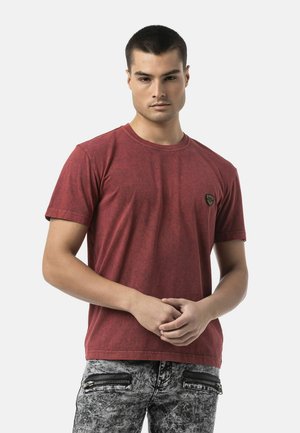 Young man in a red t-shirt and acid-washed jeans with zipper pockets standing with hands loosely clasped against a plain light background.
