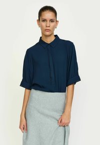 Marineblauwe blouse met korte, opgeblazen mouwen, een knoopsluiting aan de voorkant en decoratieve afwerking op de mouwen, gedragen met een grijze rok.