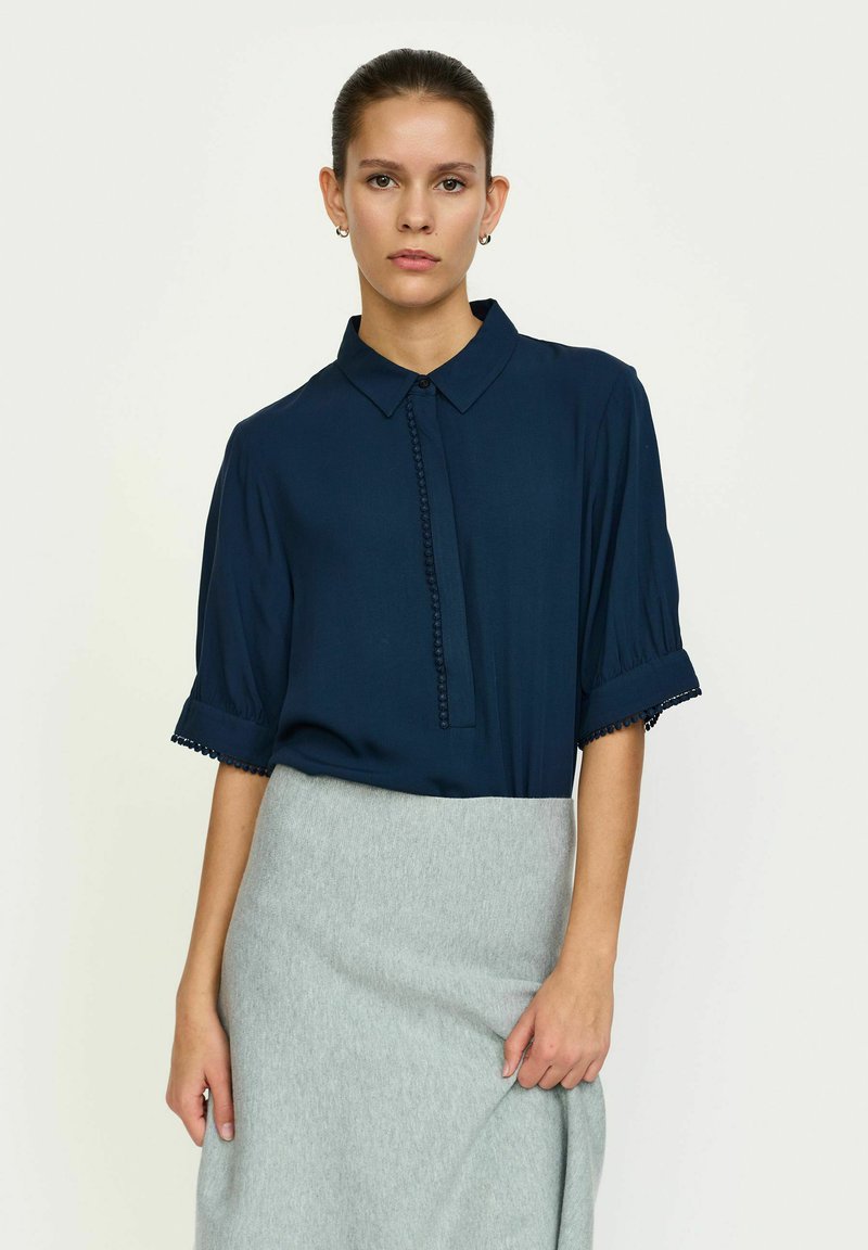 Marineblauwe blouse met korte, opgeblazen mouwen, een knoopsluiting aan de voorkant en decoratieve afwerking op de mouwen, gedragen met een grijze rok.