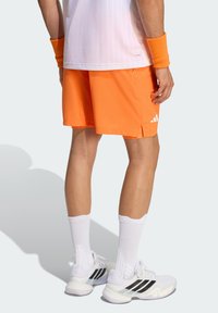 Πορτοκαλί αθλητικό σορτς με υφή, με λευκή σήμανση Adidas, συνδυασμένο με λευκές κάλτσες και παπούτσια με μαύρες λεπτομέρειες.