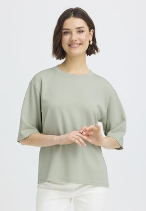 Młoda kobieta uśmiechająca się, ubrana w jasnorozielony, oversize'owy T-shirt i białe spodnie, delikatnie splecione ręce na przodzie, szare tło.