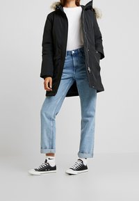 Black parka jacket with faux fur hood, zip closure, and two front pockets, paired with light blue jeans and black sneakers.