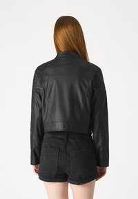 Giacca cropped in pelle nera con un design strutturato, dettagli trapuntati sulle spalle e colletto inclinato. Indossata con pantaloni corti in denim neri.