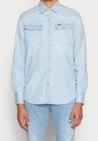 Jasnoniebieska koszula dżinsowa z długimi rękawami, ostrym kołnierzykiem, dwoma kieszeniami na piersi i metalicznymi guzikami. Charakteryzuje się zaokrąglonym dołem i luźnym krojem.