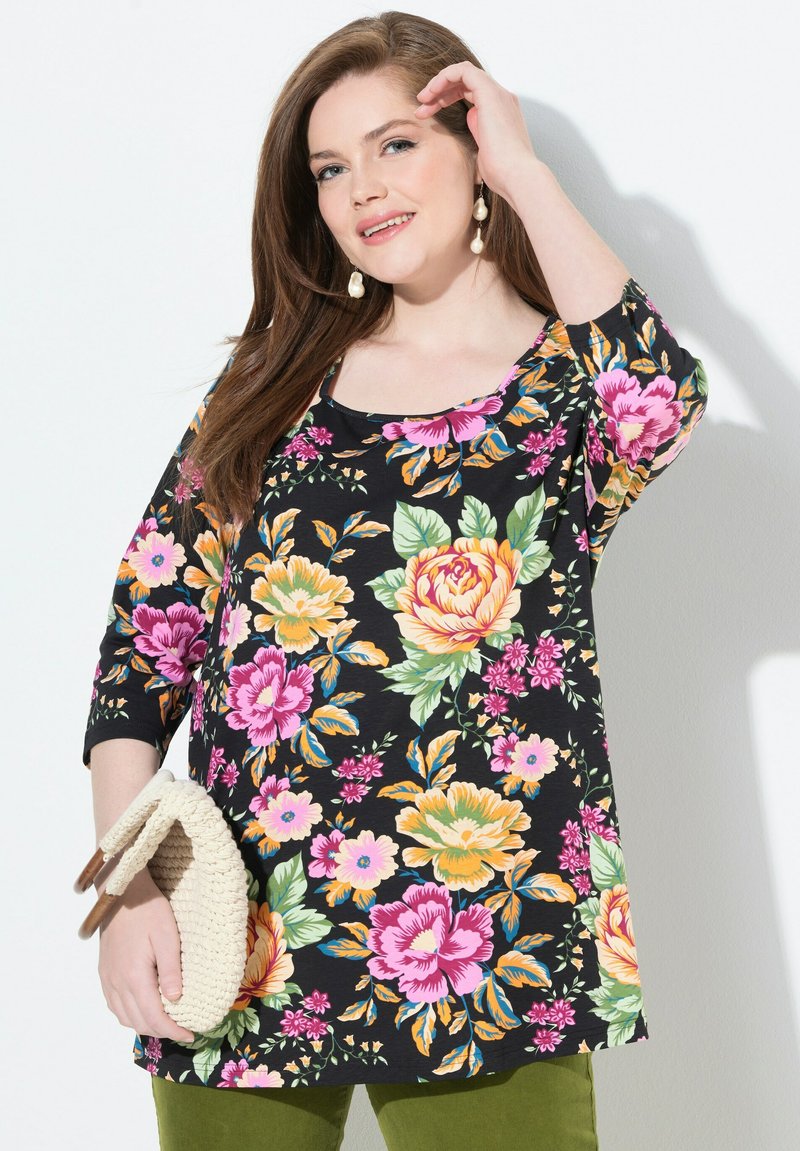 Schwarze Blumenbluse mit 3/4-Ärmeln, die mit rosa, gelben und grünen Blumen verziert ist. Dazu eine aus geflochtenem cremefarbenem Handtasche.