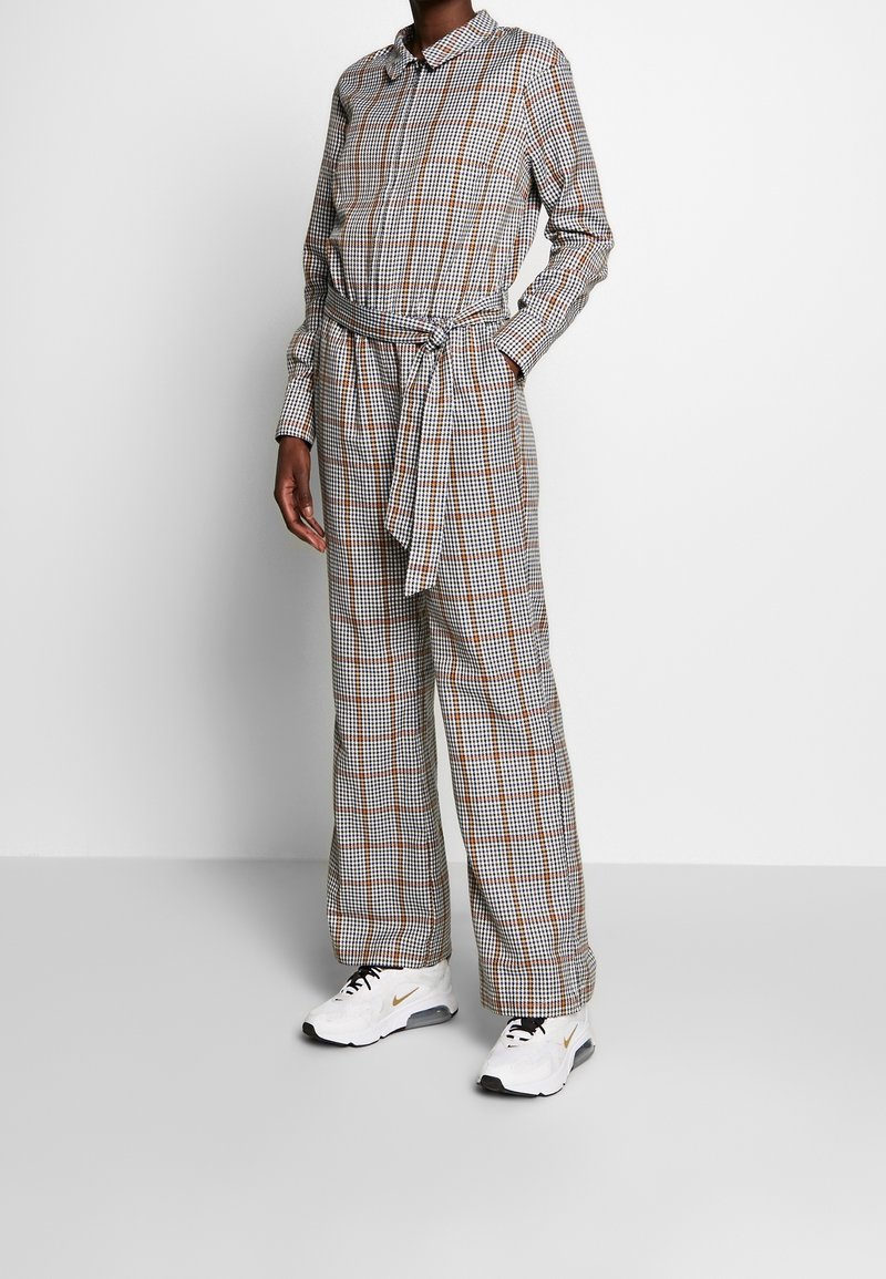 Personne portant une combinaison à manches longues à carreaux avec une ceinture à la taille et des baskets blanches avec des lacets noirs et des accents dorés.