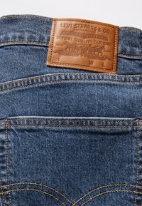 Jeans din denim albastru au o etichetă din piele maro în spate, cu text embosat și un design cu nituri. Cusăturile duble adaugă durabilitate.