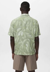 Camisa verde de manga corta con un patrón fluido en tonos verde claro, hecha de un tejido ligero. Combinada con pantalones cortos beiges.