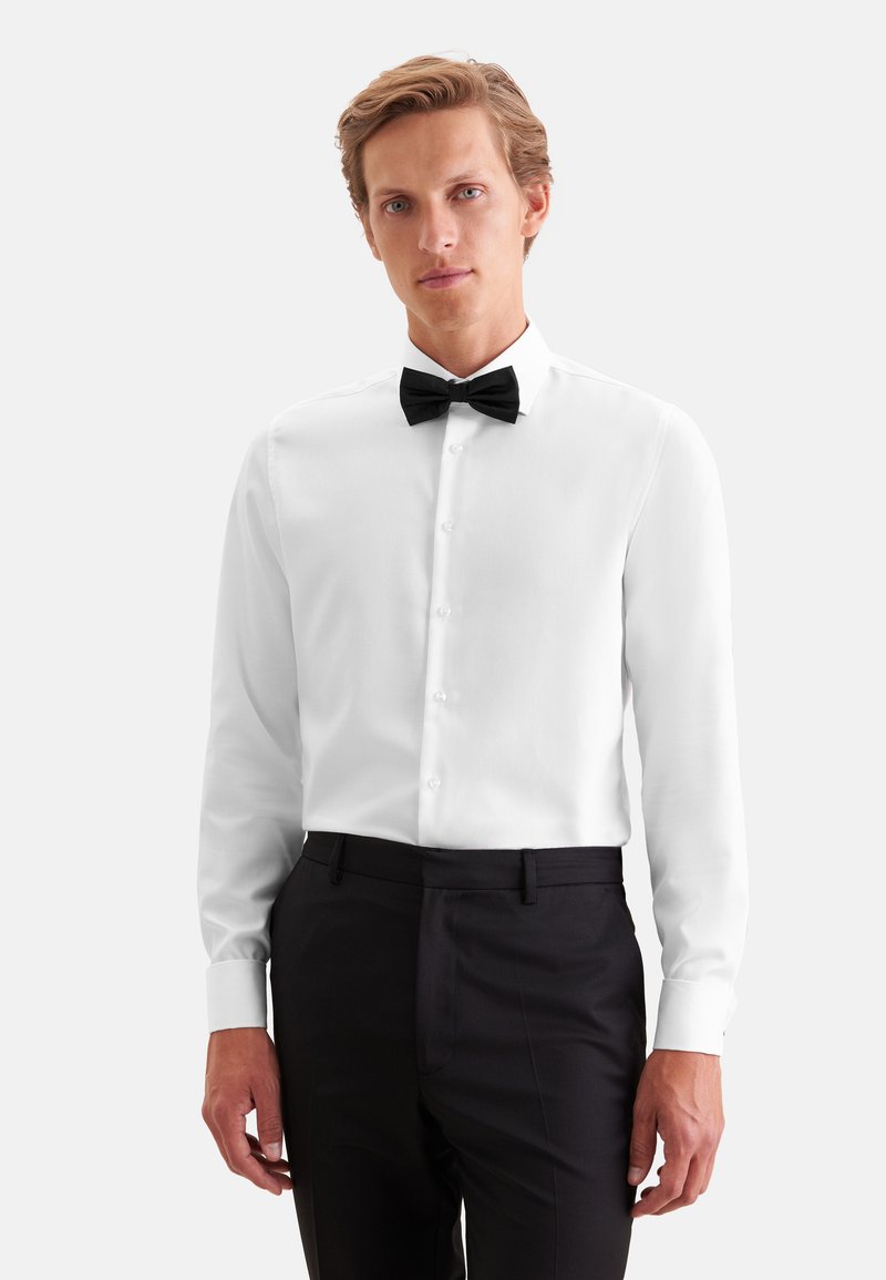 Homme portant une chemise blanche avec un nœud papillon noir et un pantalon de costume noir, debout devant un fond clair uni.