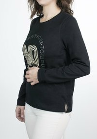 Camisa negra de manga larga con cuello redondo, que presenta un diseño gráfico texturizado en dorado y aberturas laterales. Tejido suave y elástico.