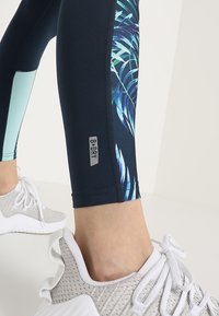 Leggings navy con pannello a stampa tropicale, realizzati in materiale elasticizzato con etichetta "B-DRY". Abbinati a scarpe da ginnastica grigie chiare.