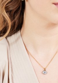 Gouden ketting met een oogvormige hanger, versierd met blauwe en heldere stenen, weergegeven tegen een lichtgekleurde kledingstof.