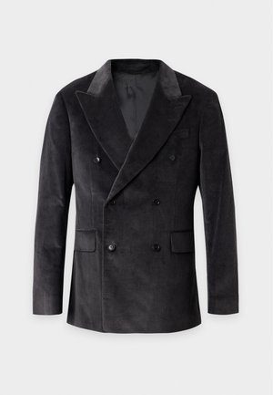 Junger Mann steht und trägt einen schwarzen zweireihigen Blazer, einen dunkelgrünen Rollkragenpullover, schwarze Hosen und schwarze Sneaker mit weißen Sohlen.