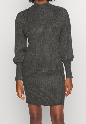 Femme portant une robe pull côtelée gris foncé avec des manches longues bouffantes et un col montant, debout devant un fond uni.