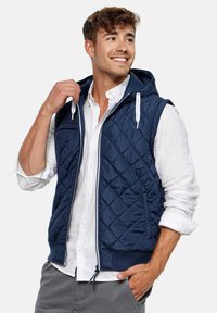 Chaleco acolchado azul marino con capucha, que presenta un cierre completo, cordones blancos y un dobladillo acanalado. Llevado sobre una camisa blanca de botones.