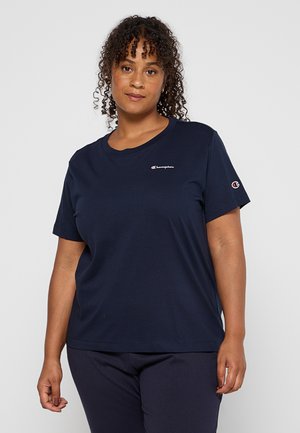 Donna con capelli ricci che indossa una maglietta Champion blu navy e pantaloni abbinati, in piedi davanti a uno sfondo chiaro e uniforme.