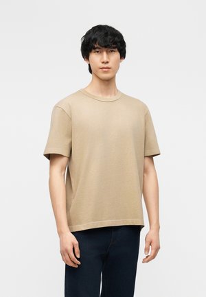 Jeune homme aux cheveux noirs portant un t-shirt beige uni à manches courtes et un jean bleu foncé, debout devant un fond blanc.