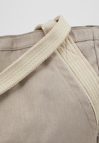 Gros plan sur un coin de sac fourre-tout en toile beige avec des anses cousues en tissu assorti qui se croisent au niveau du bord supérieur.