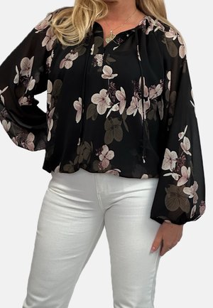 Vrouw die een zwarte doorschijnende blouse draagt met roze en bruine bloemenpatronen en een witte broek, met één hand op haar heup.