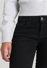 Černé džíny z denimu s úzkým střihem, se přední kapsou, zapínáním na knoflík a matným povrchem, doplněné šedým svetrem.