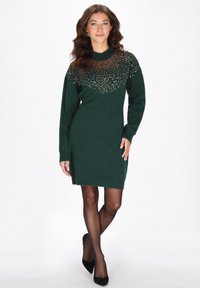 Sukienka dzianinowa w głębokiej zieleni z fakturowaną, zdobioną górną częścią. Długie rękawy i boczny rozporek, uzupełnione czarnymi szpilkami.