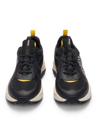 Paire de sneakers sportives noires avec des semelles beiges, des languettes jaunes et le logo "HUGO" sur la languette et le côté, vues de face.