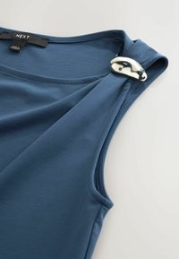 Haut bleu sans manches avec une boucle décorative ovale métallique sur l'épaule gauche et une étiquette "NEXT" près de l'encolure.