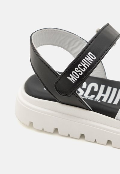 Sandalias sintéticas negras con una suela de plataforma blanca, correa ajustable y la marca "MOSCHINO" en el exterior de la correa.
