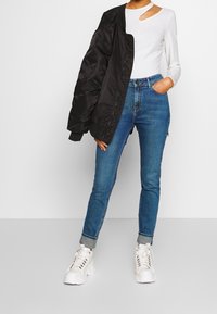 Chaqueta bomber negra sobre una camiseta de manga larga blanca con detalle de corte, combinada con jeans ajustados azules y zapatillas blancas gruesas.