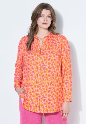 Kobieta z luźnymi, falującymi brązowymi włosami, ubrana w jaskrawą pomarańczowo-różową bluzkę w panterkę oraz różowe spodnie, stojąca z jedną ręką w kieszeni.