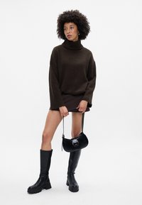 Pārmērīga tumši brūna džemperis ar augstu apkakli un ribotu faktūru. Modelis valkā plisētu mini svārku, melnus līdz ceļiem zābakus un spīdīgu rokassomu.