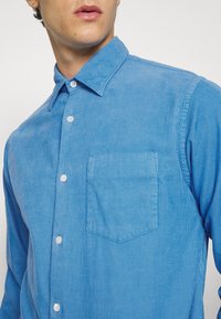 Camicia di corduroy azzurra chiara con colletto button-down, maniche lunghe, taschino sul petto e sottili righe verticali. I bottoni sono bianchi.