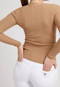 Světle hnědý pletený svetr s texturovaným kabelkovým vzorem, přiléhavý silueta a dlouhé rukávy, kombinovaný s bílými džíny s logem a zadními kapsami.
