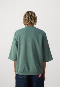Løs-fit grøn sweatshirt lavet af blødt stof, med trekvartærmer, rund hals og et lille logomærke på venstre side af hoften.