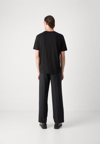 Zwart t-shirt met korte mouwen van katoen, ronde halslijn, gecombineerd met loszittende donkere broeken en zwarte schoenen, weergegeven vanuit de rug.