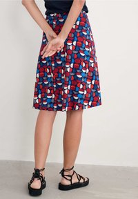 Personne debout, les mains jointes derrière le dos, portant une jupe jusqu'aux genoux avec des motifs géométriques rouges, bleus et blancs, ainsi que des sandales noires à brides.