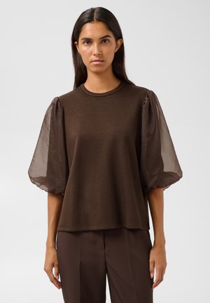 Femme aux longs cheveux foncés portant un haut marron avec des manches bouffantes transparentes et un pantalon marron assorti, debout devant un fond uni.