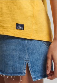 T-shirt di cotone giallo con un piccolo etichetta a logo blu navy sull'orlo, abbinato a una gonna di jeans blu con orlo grezzo e dettaglio spacco.