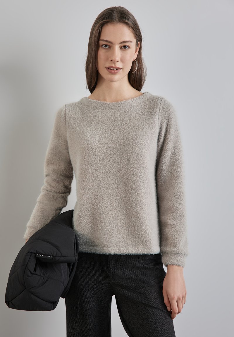 Suéter de color gris claro y suave con un escote redondeado y mangas largas, combinado con una chaqueta acolchada negra, llevada sobre pantalones oscuros.