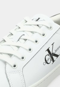 White sneaker featuring a smooth leather exterior, round toe, flat cotton laces, and a black "CK" logo on the side.