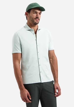 Man draagt een lichtgroen, kort mouwig overhemd met knopen, donkere broek en groene pet, staand tegen een witte achtergrond.