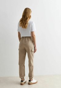 Pantaloni cargo beige con vita elastica e polsini alla caviglia, dotati di tasche laterali. Indossati con una maglietta bianca a maniche corte e sneakers.