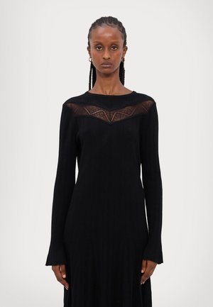 Rochie tricotată neagră cu mâneci lungi, având un decolteu din dantelă transparentă. Poală evazată cu manșete delicate cu franjuri. Design simplu și elegant.
