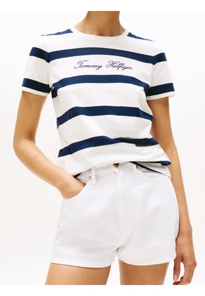 T-shirt di cotone blu navy e bianca a righe, con "Tommy Hilfiger" ricamato in corsivo, abbinata a pantaloncini di denim bianchi, mani nelle tasche.