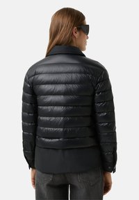 Schwarze Steppjacke mit glänzendem Finish, horizontalen Nähten, Kragen und leicht verlängertem Saum. Kombiniert mit dunkelgrauen Jeans.