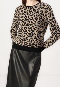 Kobieta w beżowo-czarnym swetrze w panterkę, zestawionym z czarną sk�órzaną spódnicą, na jednolitym białym tle.