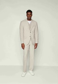 Beige suit made of smooth fabric, featuring a single-button jacket, slim-fit trousers, and a light grey vest underneath. White sneakers worn.