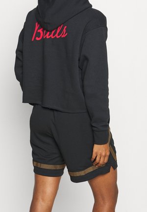 Person trägt schwarzen, kurz geschnittenen Hoodie mit rotem "Bulls"-Aufdruck auf dem Rücken und schwarze Shorts mit goldenen Mesh-Streifen an den Seiten und am Saum.